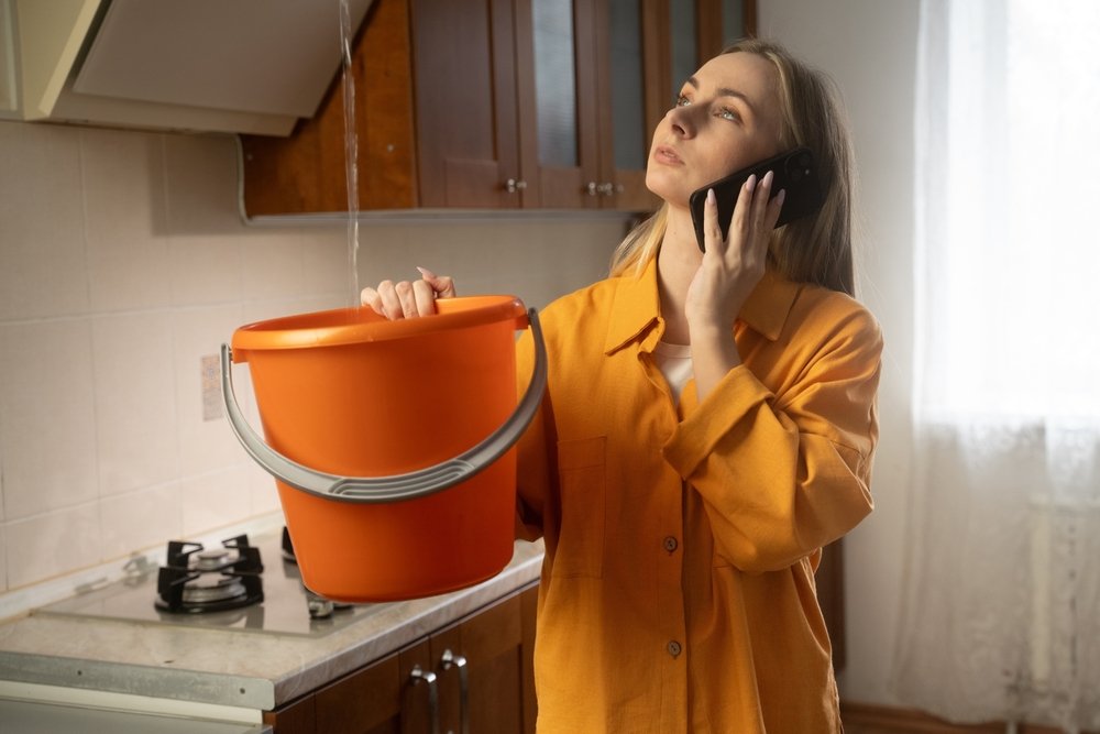 Worried woman collecting dripping water in orange bucket while calling plumber or insurance company to report ceiling water leak in kitchen