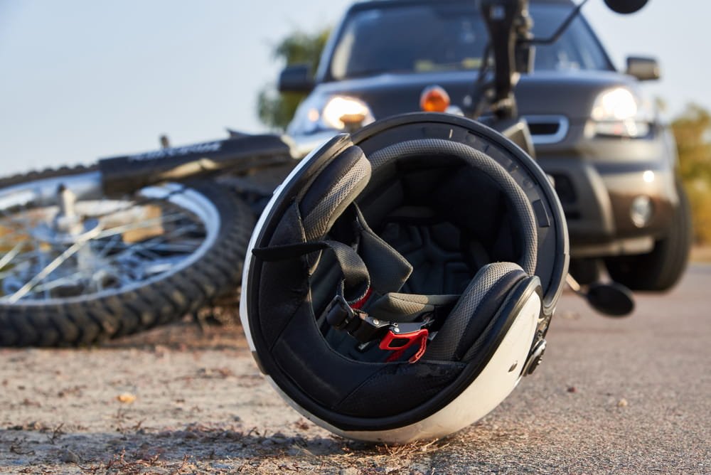 Casco de motocicleta en primer plano con un auto al fondo tras un accidente, ilustrando cómo se atribuye la culpa al motociclista en Florida